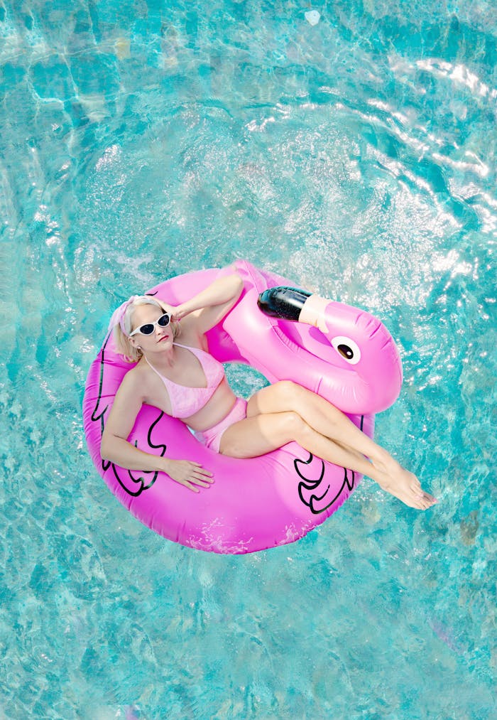 A woman in sunglasses lounges on a pink flamingo float in a bright blue swimming pool.